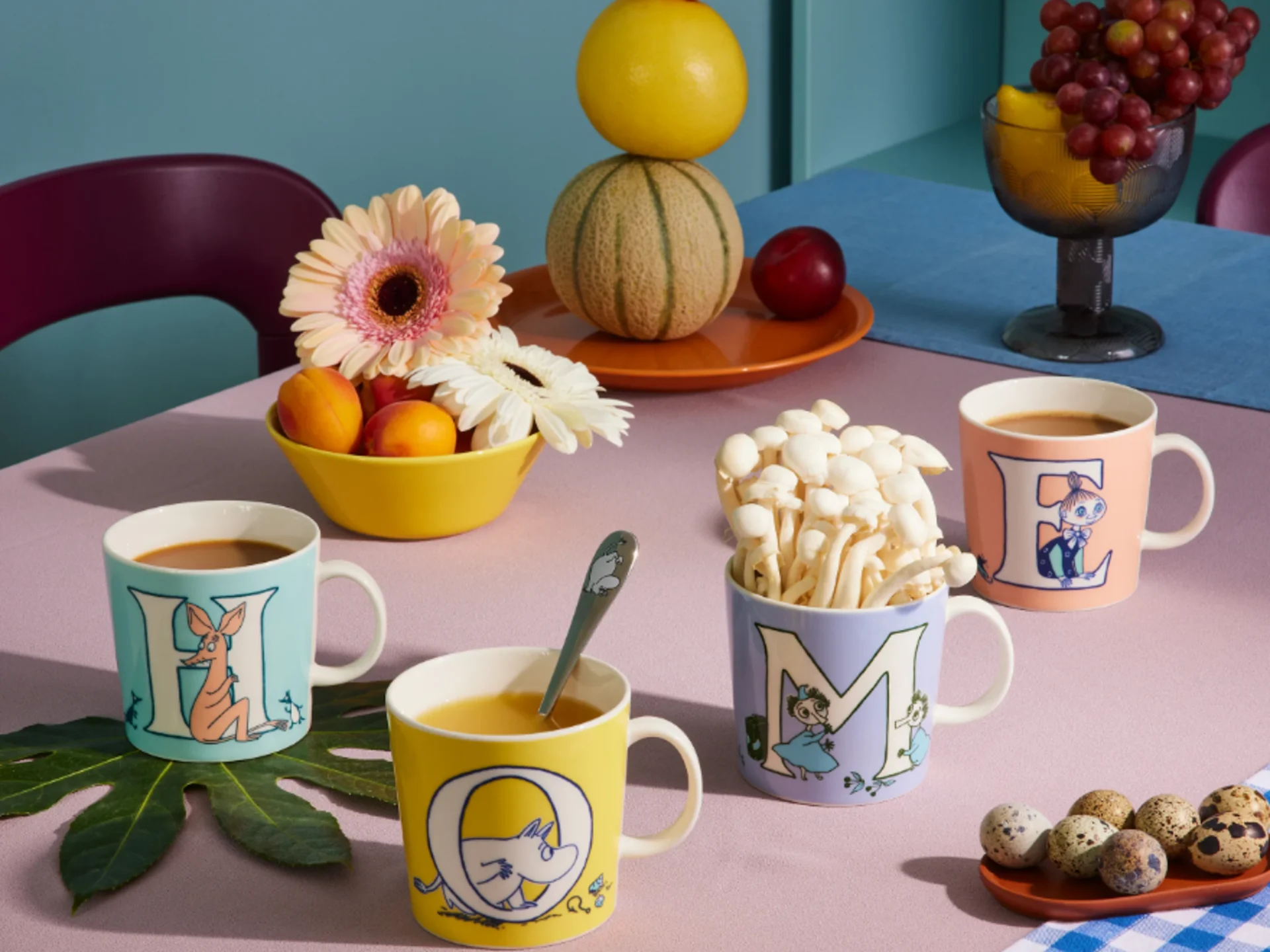 H、O、M、Eの文字が描かれた4つのカラフルなムーミンマグカップ。コーヒーとキノコが入り、果物や花と共にテーブルに置かれている。