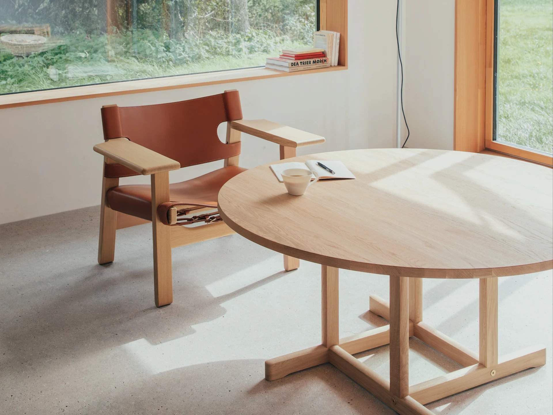 明るい部屋に茶色の革製アームチェアと、カップとノートが置かれた丸い木製テーブルがあり、窓から緑が見える。