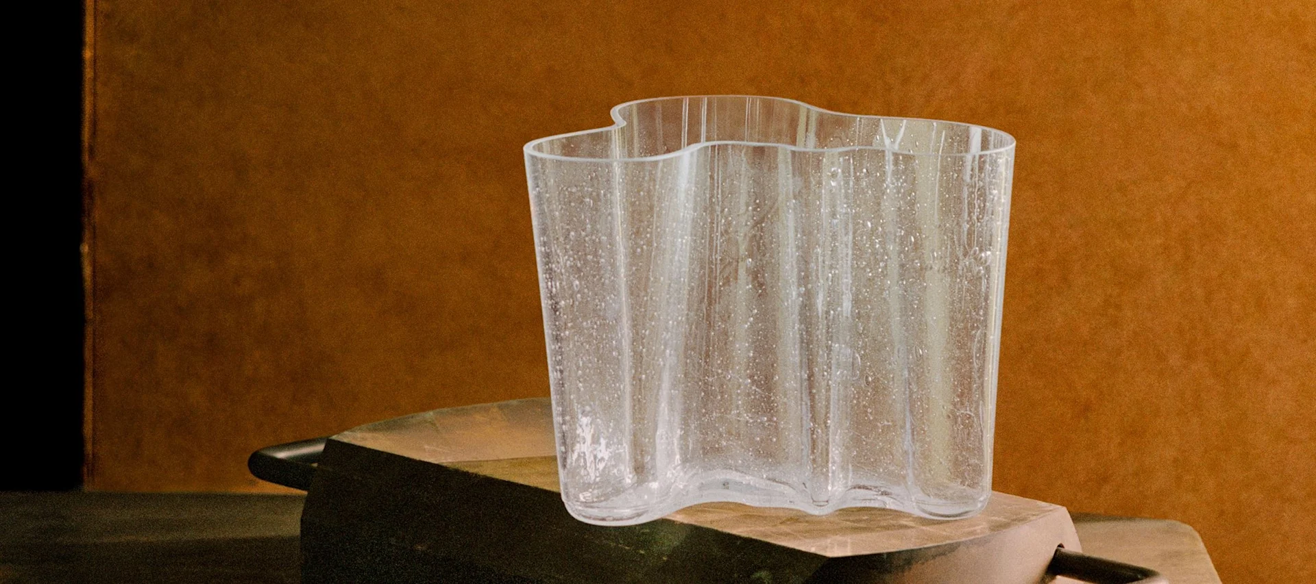 波状の透明なガラスの花瓶が、テクスチャのある斑点とともに、素朴な暗い表面の上に暖かい茶色の背景を背にして置かれている。