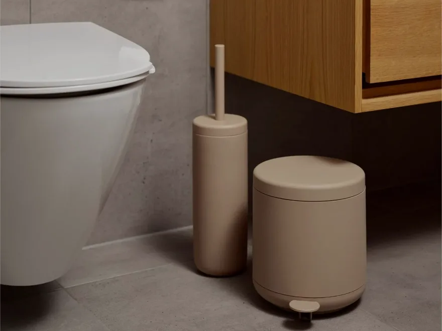 白いトイレ、ベージュのトイレブラシホルダーとペダルビンがタイル張りの床に置かれたモダンなバスルーム。