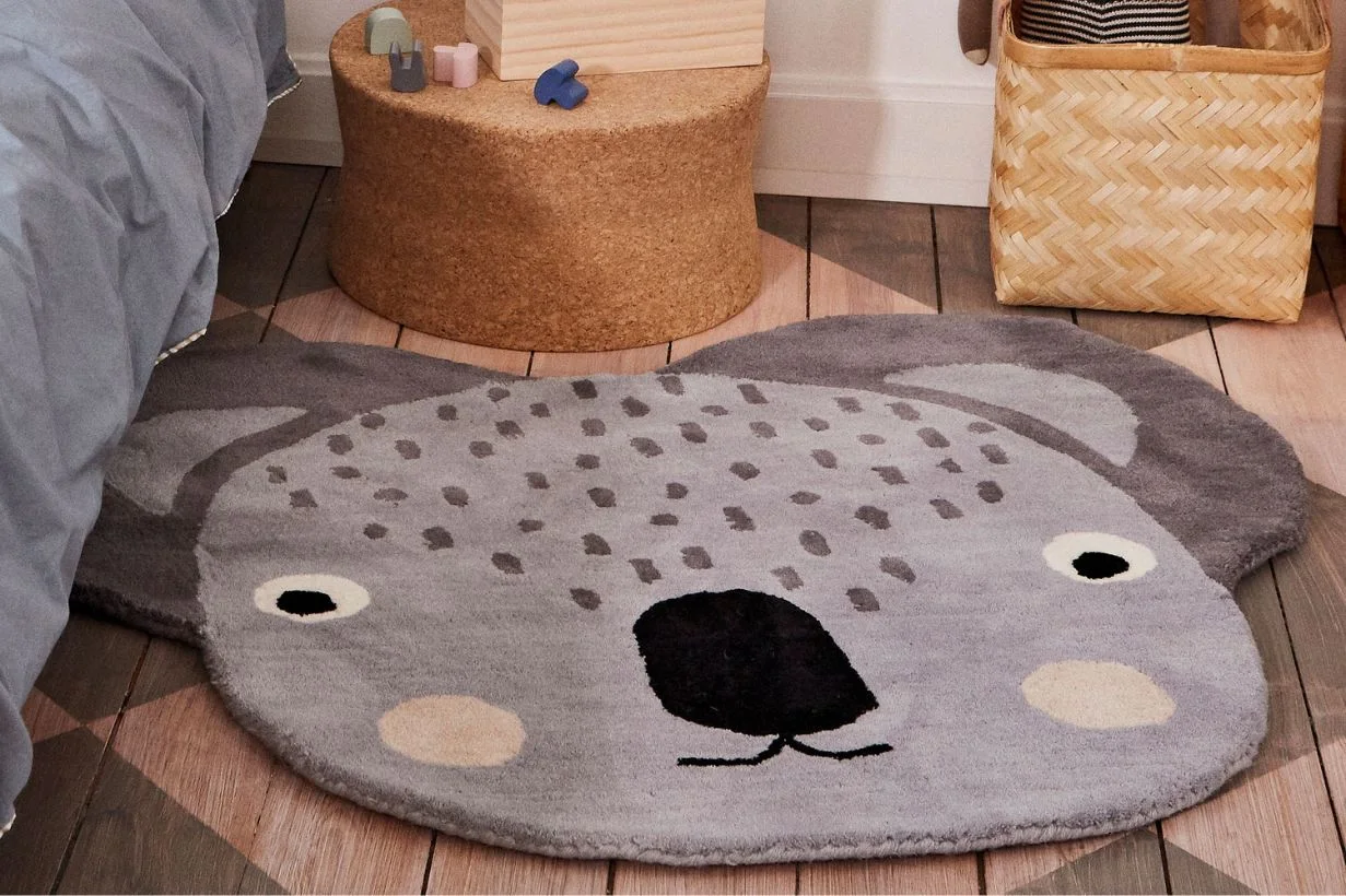黒い鼻と目、薄いベージュの頬を持つ、グレーのコアラ型ラグが子供部屋の木の床に敷かれている。