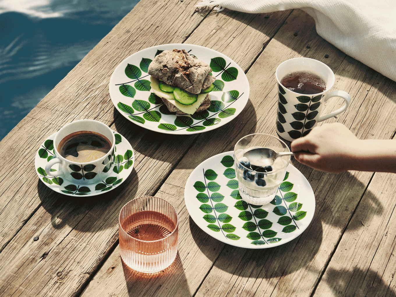木製の桟橋での朝食：葉模様の皿にはサンドイッチ、コーヒー、紅茶、ブルーベリーヨーグルト、ピンクの飲み物があります。