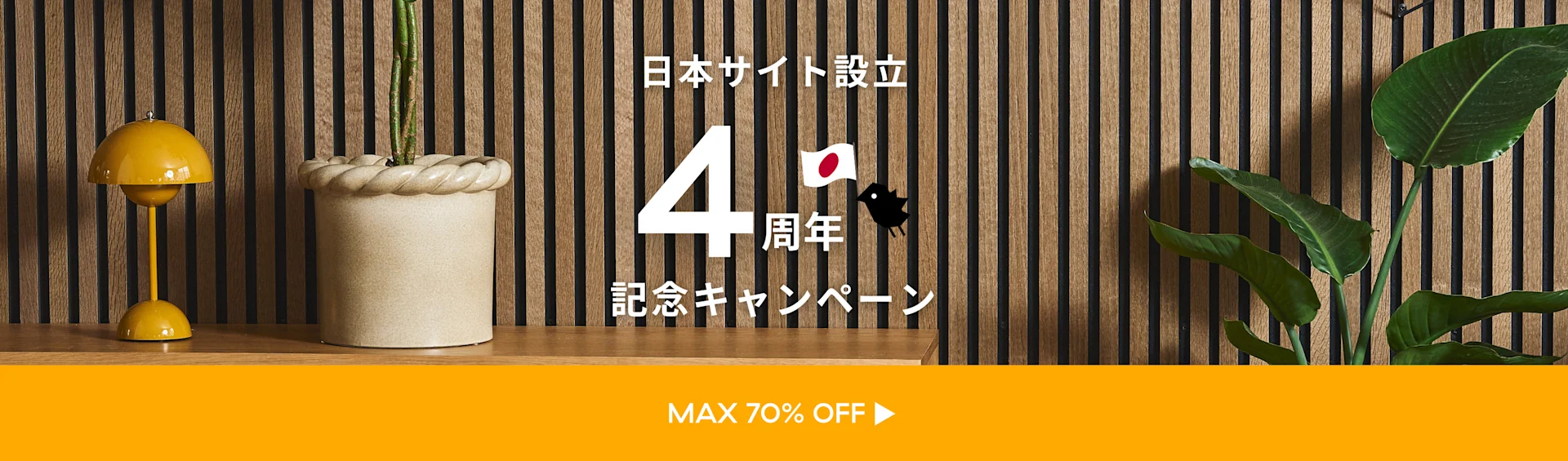 日本サイト開設4周年記念キャンペーンのバナー。黄色のランプ、鉢植え、観葉植物が木製スラット壁を背景に配置され、「MAX 70% OFF」と表示されています。