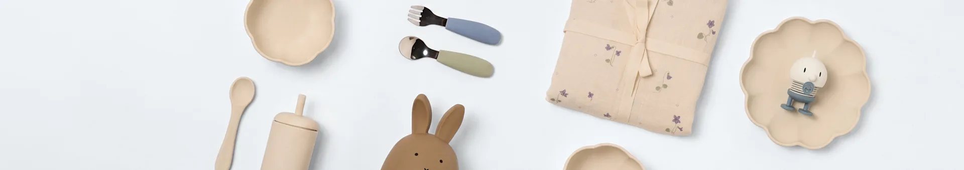 ベビー用食器と遊び道具の平置き：ベージュのボウル、青いフォーク、緑のスプーン、花柄のブランケット、皿の上のキャラクターキャンドル、木のスプーン、うさぎのおもちゃ。