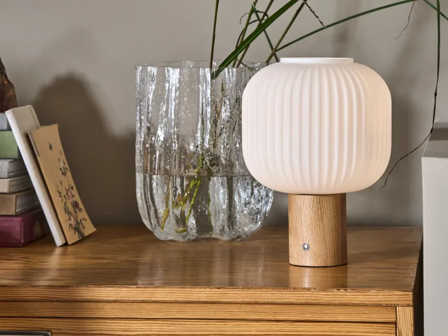 白いリブ付きテーブルランプ、明るい木製ベース、木製表面に置かれた緑の植物が入ったテクスチャードガラスの花瓶の隣。