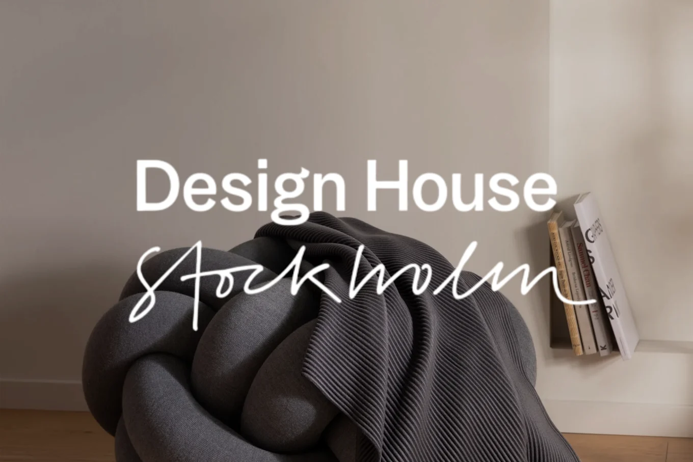 グレーの結び目型プーフ、リブ編みのスロー、本の山、そして「Design House Stockholm」の文字があるインテリア。