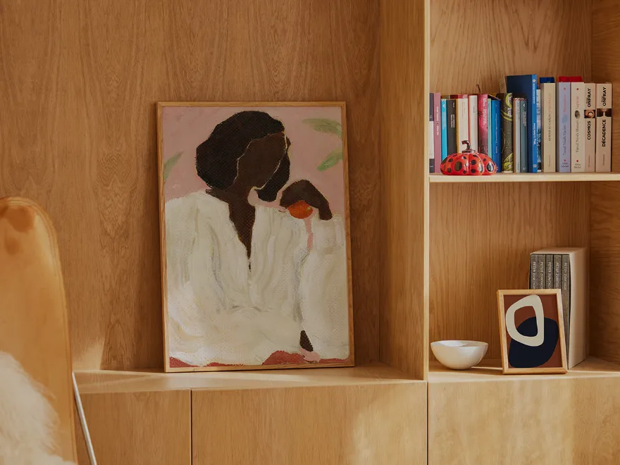 ピンクの背景にオレンジを持つ人物の絵画、木製棚に本や装飾品と一緒に飾られている。