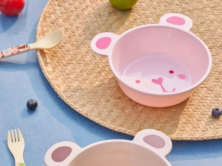 クマの形をしたボウル2つ、ピンクとベージュ、お揃いのスプーンとフォーク、フルーツと一緒に編みマットの上。