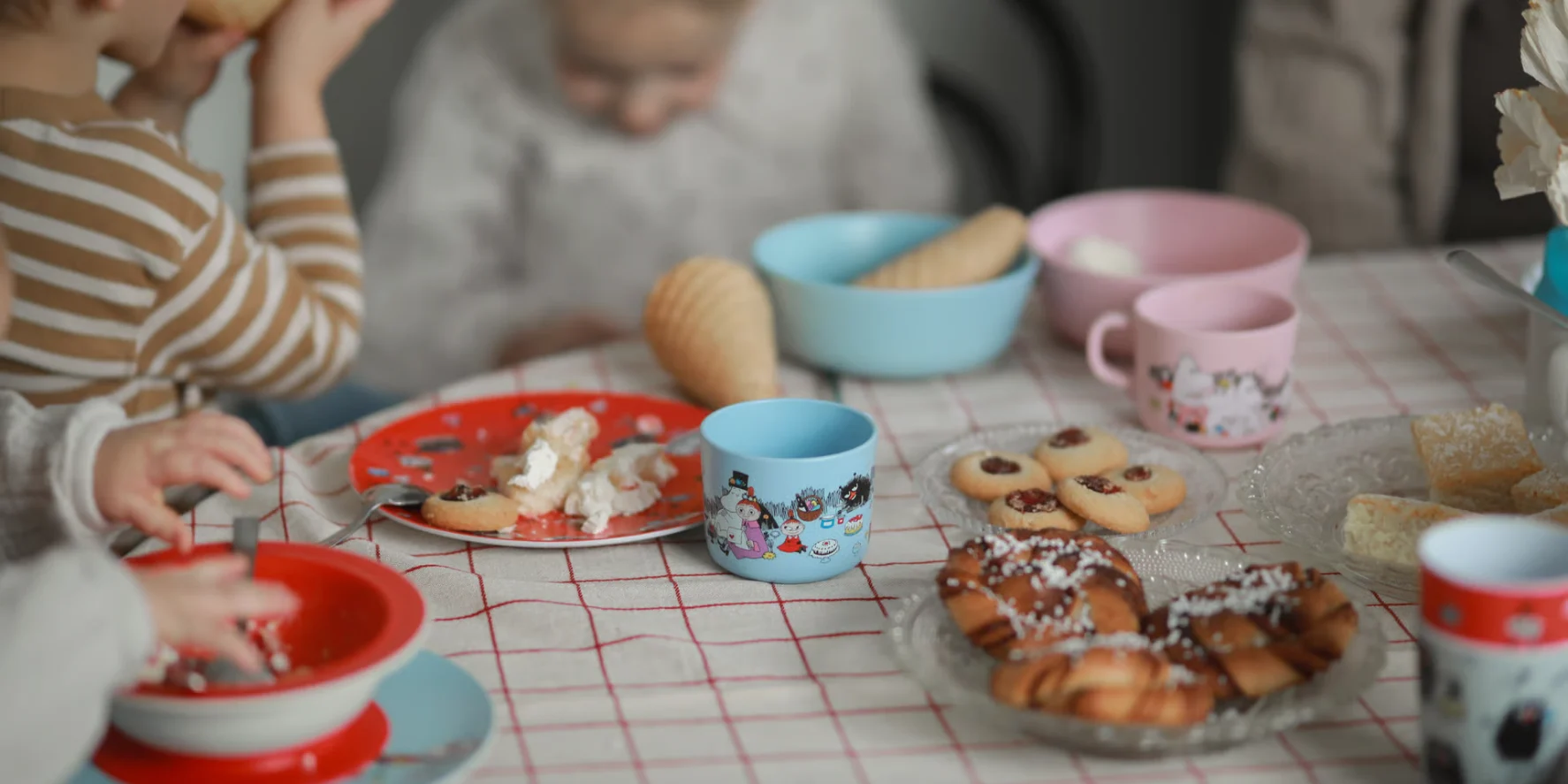 カラフルな食器が並ぶテーブルで、子供たちが様々なパンやデザートに手を伸ばしている様子。