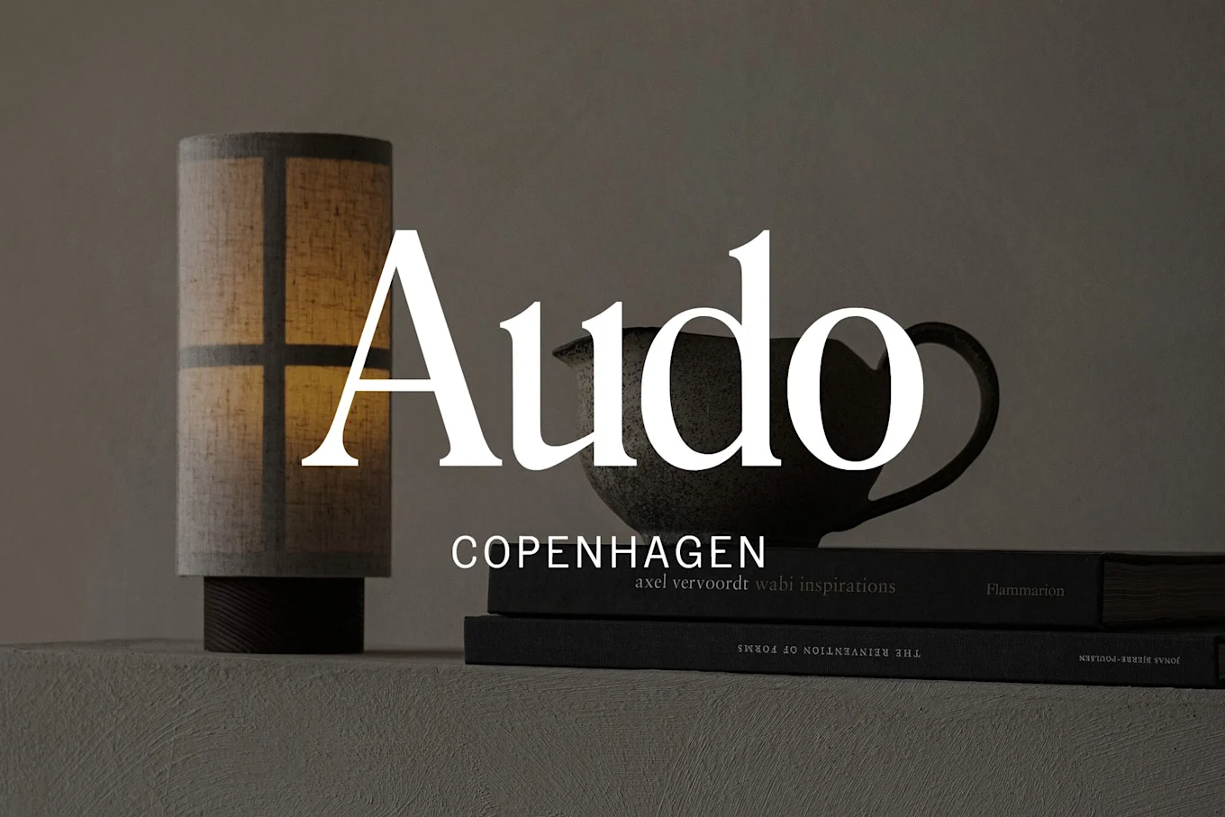 薄暗いシーンに、ランプ、暗いマグカップ、積み重ねられた本。'Audo Copenhagen'の文字が重ねられています。