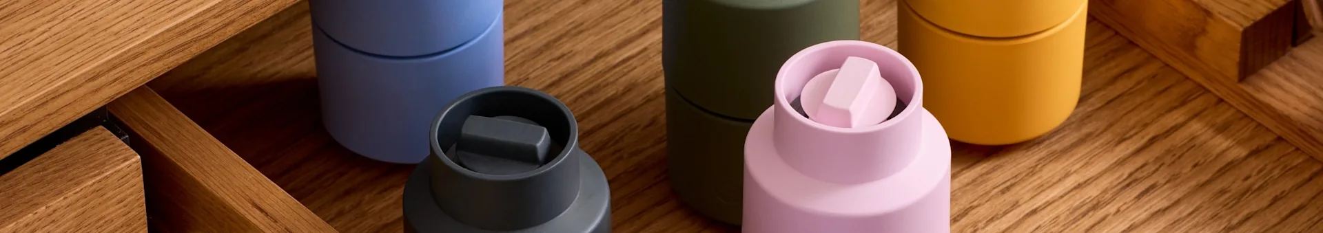 5つのカラフルな容器：ダークグレー、ライトブルー、オリーブ、特徴的なキャップ付きのライトピンク、マスタードイエロー、木製の上。