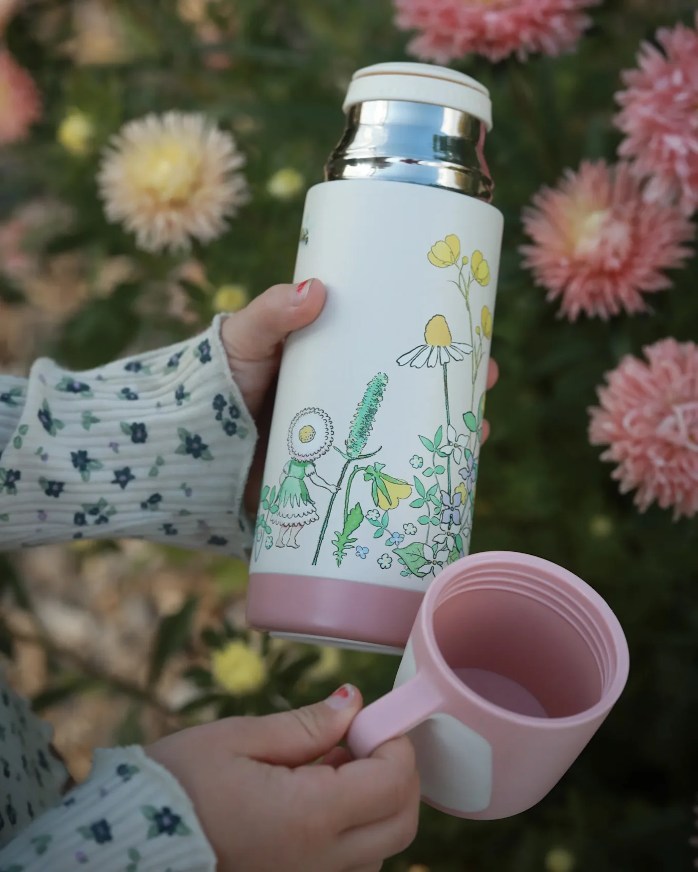 子供の手が白い水筒をピンクのカップと並んで持ち、水筒にはデイジーの頭の少女と花柄が描かれ、背景はぼやけた花々。
