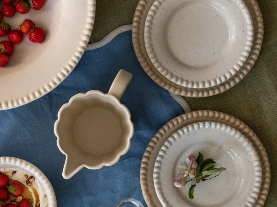 斑点のあるセラミック皿、イチゴのボウル、クリーマーが青と緑のリネンテーブルクロスに置かれ、リンゴの花が添えられています。