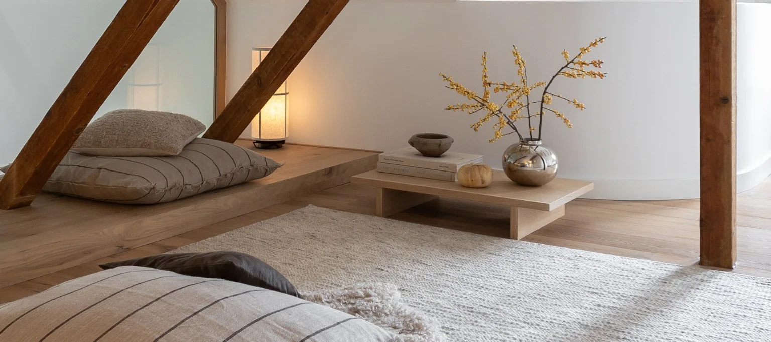 明るい木の床と白い壁の居心地の良い部屋で、フロアクッション、光るランプ、黄色の実の入った銀色の花瓶が置かれた低いテーブルがあります。