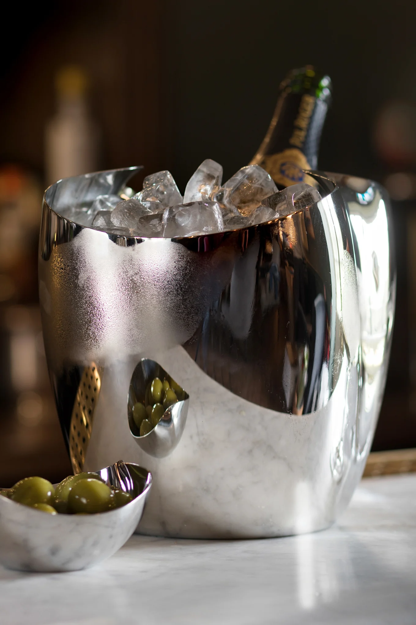 シャンパンボトルと氷が入った反射する銀色のワインクーラー。横には緑のオリーブが入った小さなボウルが2つ大理石の上にあります。