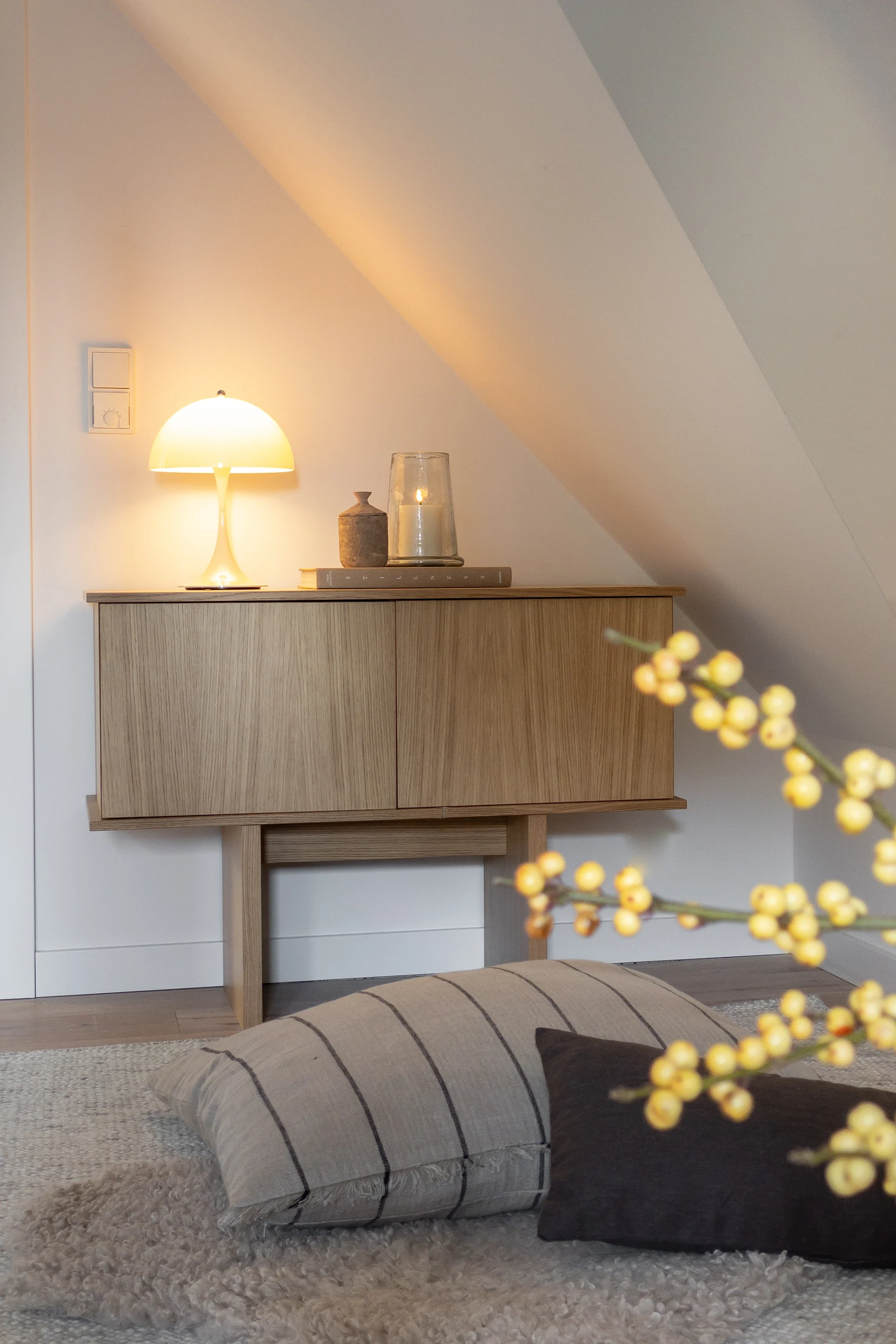 温かい光を放つキノコ型ランプと灯されたキャンドルが置かれた木製キャビネットが、白い傾斜のある壁の居心地の良い部屋に配置されている。