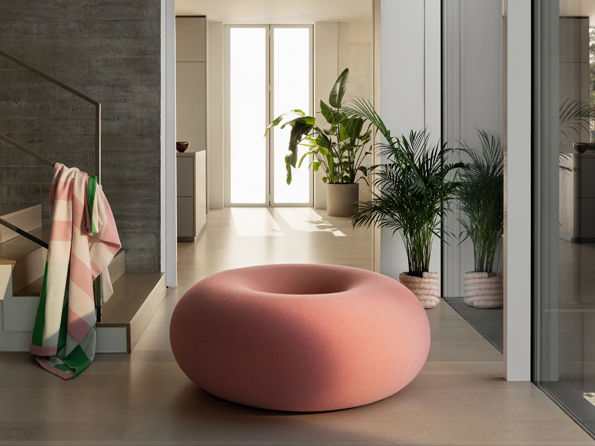 ドーナツ型の大きなピンクのプーフが、植物とコンクリートの壁があるモダンな部屋の明るい木製の床に置かれています。