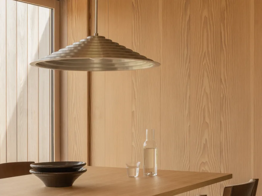 真鍮色の段状円錐形ペンダントライトが、軽い木製のテーブルの上、積まれた濃い陶器のボウル、水筒、グラスの上を照らしている。