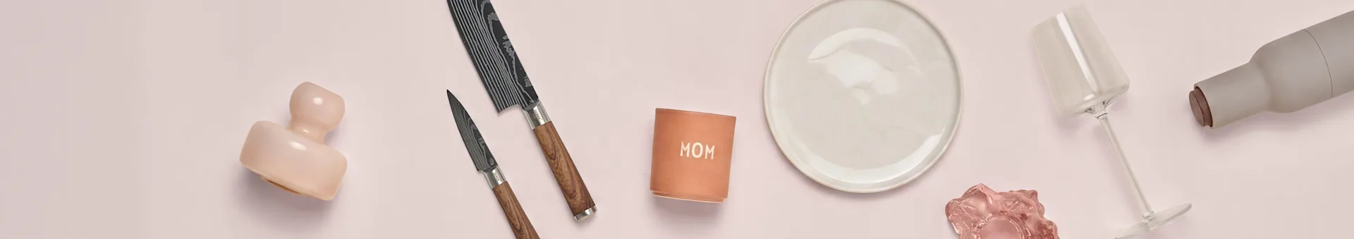 ピンクのセラミック、2本のナイフ、「MOM」カップ、白い皿、ピンクのグラス、ワイングラス、灰色のボトルがピンクの表面に置かれています。