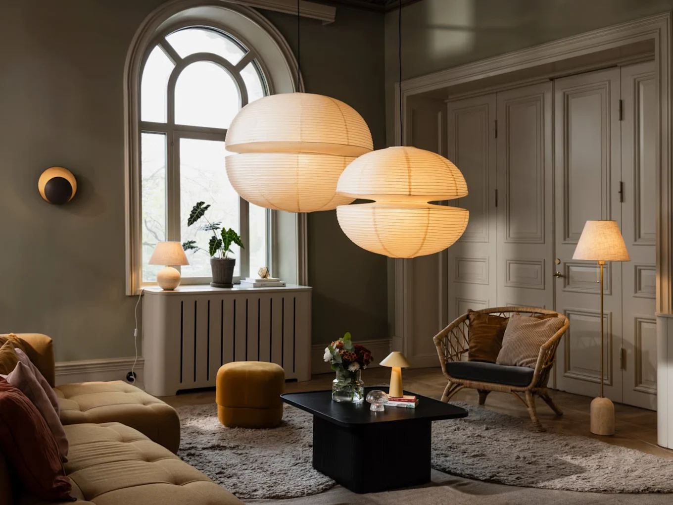 2つの大きな紙製ペンダントライト、黄色いソファ、籐椅子、様々なランプがある居心地の良いリビングルーム。
