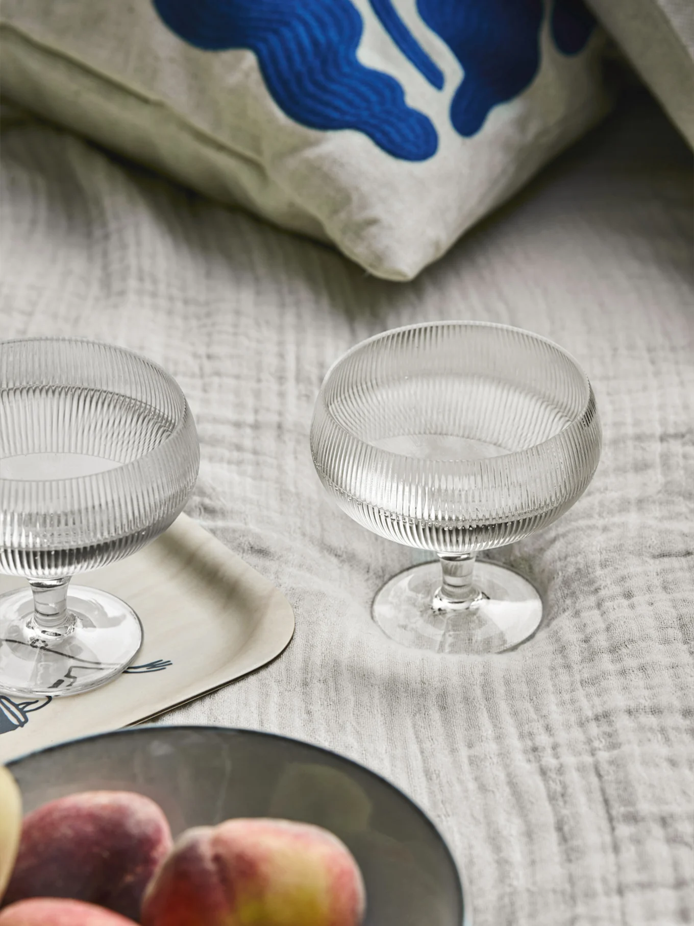 2つのリブ付きガラスデザートボウル、青い抽象画の枕、木製トレイ、明るい布地の上の桃。