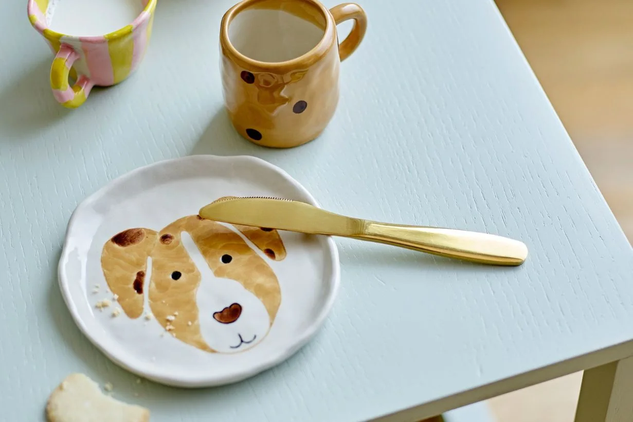 茶色と白の犬の顔が描かれた白い皿、金色のナイフ、2つのマグカップが青い木製テーブルに置かれている。
